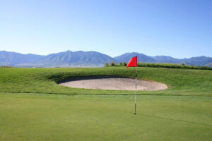 Flag pin and green in front of a sand bunker with mountains in the background.